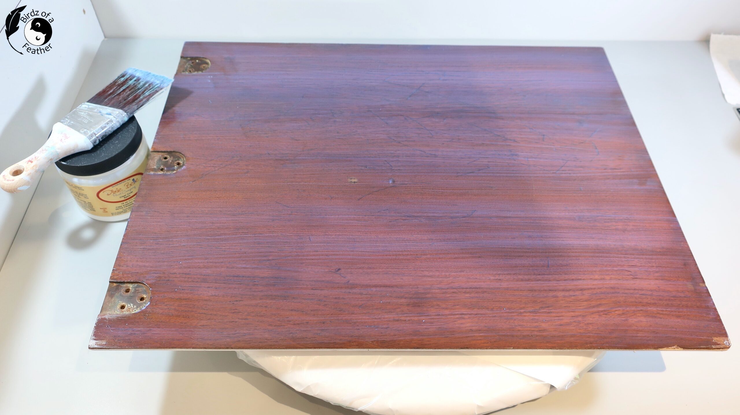 Jar of clear coat with brush resting on top beside wood sewing table lid that has been clear coated