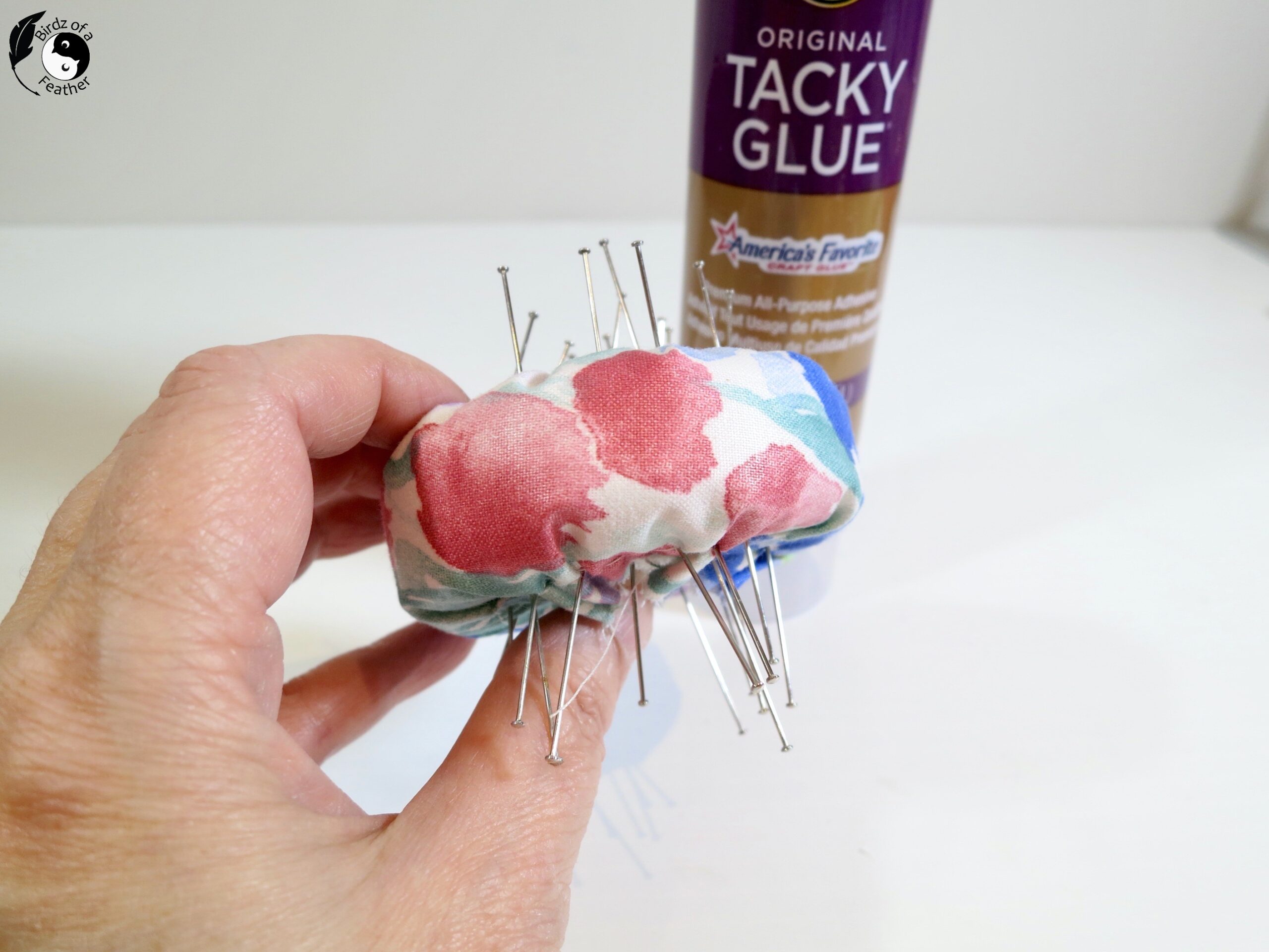 Hand showing both sides of fabric wrapped foam as it dries with pins sticking out and bottle of tacky glue in background