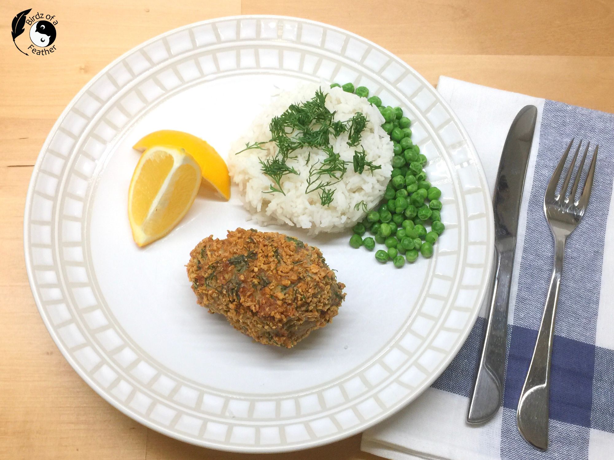 One chicken Kyiv on a pleat served with lemon wedges, rice and peas, beside a blue and white napkin, knife, and fork