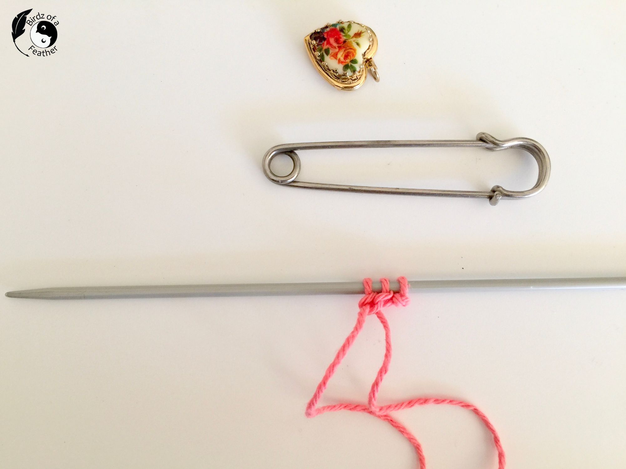 3 stitches cast on a knitting needle with kilt pin and heart locket in background
